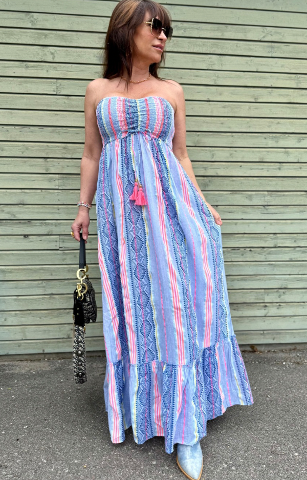 Axelbandslös lång klänning i jeansblått och coral med tofsar under bysten. Tillverkad i 100% bomull. One size passar XS till M. Modellen har bystmått 93 cm.