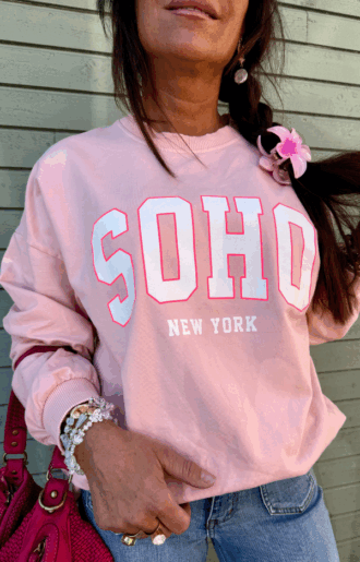 Collegetröja i puderrosa med rosa SOHO – New York-tryck, fotad framifrån med muddar vid ärmslut och midja, i mjuk bomull.
