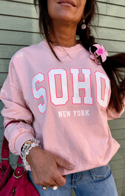Collegetröja i puderrosa med rosa SOHO – New York-tryck, fotad framifrån med muddar vid ärmslut och midja, i mjuk bomull.