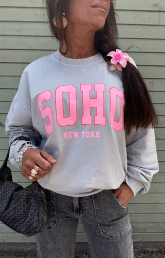 Collegetröja i bomull med rosa SOHO – New York-tryck, muddar vid ärmar och midja, fotad framifrån på galge eller modell.