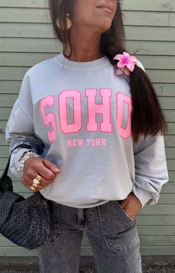 Collegetröja i bomull med rosa SOHO – New York-tryck, muddar vid ärmar och midja, fotad framifrån på galge eller modell.