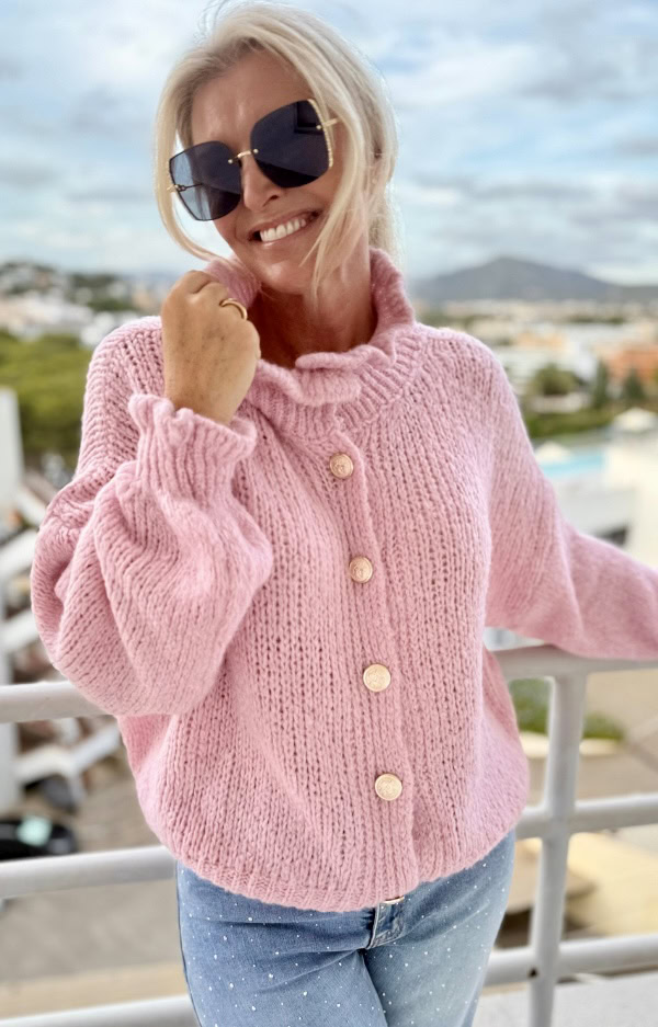 Ljusrosa stickad kofta med guldknappar och volanger vid hals och ärmslut. Mjuk, feminin och elegant design i one size (XS–L).